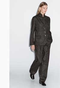 Donna che cammina in un completo elegante a righe sottili di colore scuro, con giacca a collo alto e pantaloni a gamba larga, indossando scarpe nere lucide, su uno sfondo neutro.