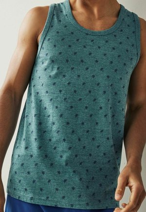 Uomo che indossa una canottiera verde senza maniche con un piccolo motivo scuro a palme e pantaloncini blu davanti a una parete chiara e liscia.