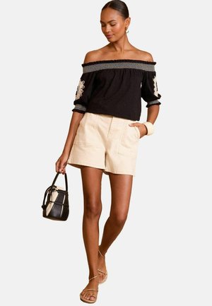 Black off-the-shoulder top with white embroidery and puffed sleeves, paired with beige shorts and strappy sandals. Small black handbag.