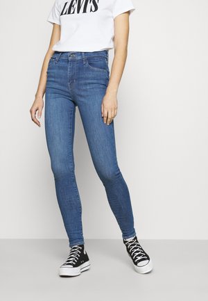 Osoba ubrana w niebieskie, dopasowane dżinsy z wysokim stanem, białą koszulkę Levi's oraz czarne sneakersy typu high-top na jednolitym jasnym tle.