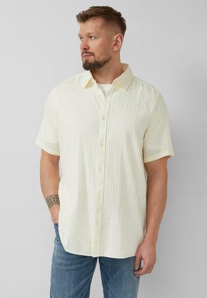 Man met baard, gekleed in een kortärmlig, gebroken wit gestreept overhemd met knopen en blauwe spijkerbroek, staand met één hand in zijn zak, kijkend naar links.