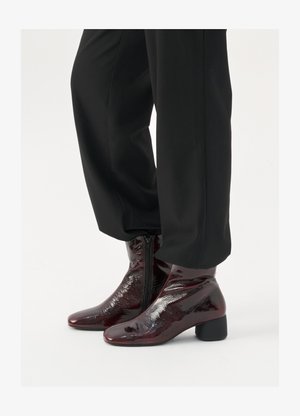 Bottines en cuir verni bordeaux avec un bout pointu, fermeture éclair sur le côté et talon épais, portées avec un pantalon noir fluide.