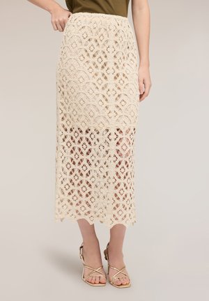 Beżowa midi spódnica z koronki szydełkowej z falistym dołem, noszona z oliwkowym topem i złotymi sandałami na wysokim obcasie na paseczkach, modelka trzyma pasek spódnicy.