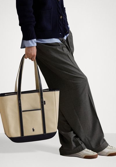 Béžová plátenná taška s tmavomodrými zvýrazneniami, pevným držadlami a predným vreckom. Obsahuje malé logo na prednej strane.