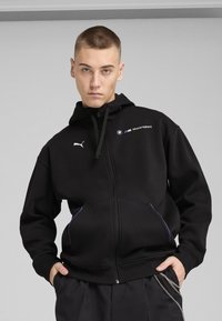 Haut de sweatshirt zippé noir en tissu doux, avec une capuche, des accents violets et des logos de BMW Motorsport et Puma sur la poitrine.
