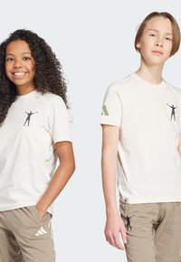 Dos niños llevan camisetas de algodón beige con un gráfico negro en el pecho. Las camisetas tienen toques verdes en las mangas.