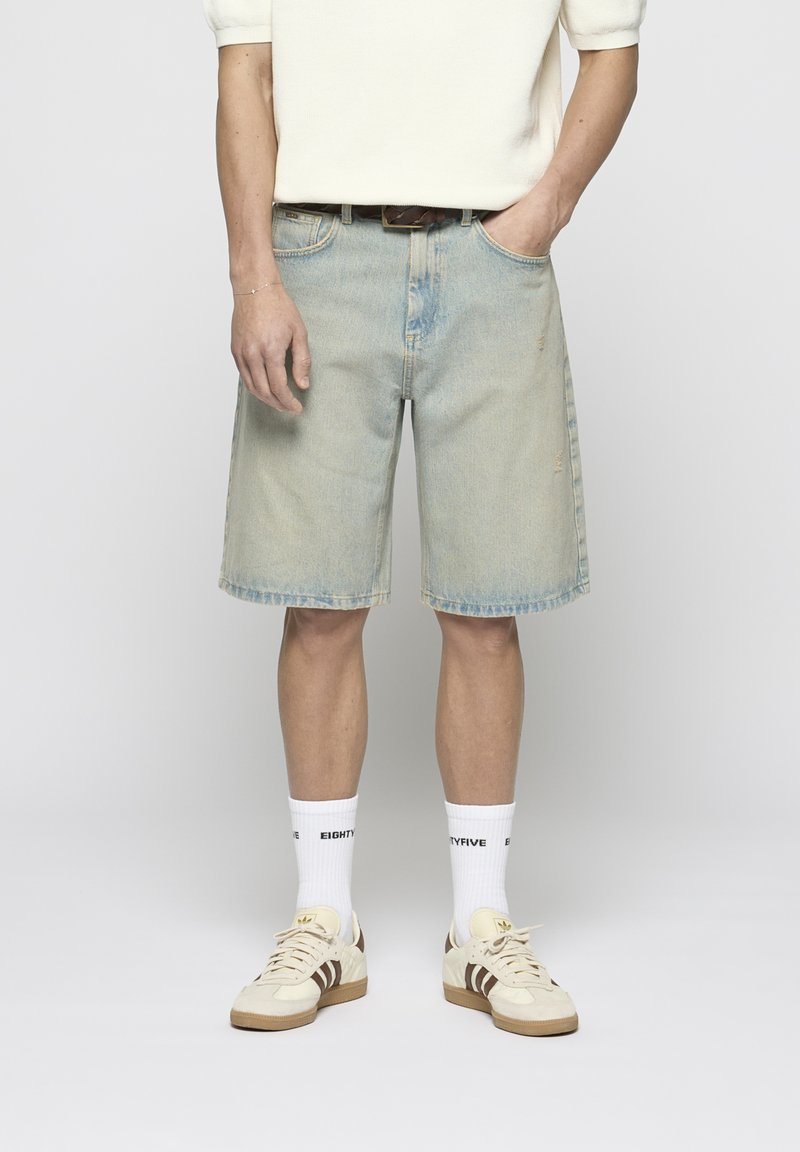 Pantalones cortos de mezclilla azul claro con un ajuste relajado, dobladillo deshilachado y diseño clásico de cinco bolsillos. Combinados con calcetines blancos y zapatillas beige.