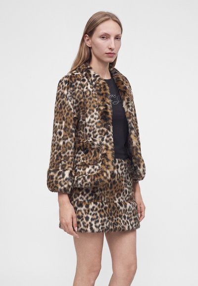 Femme portant une veste en fausse fourrure imprimé léopard et une mini-jupe assortie sur un top noir, sur fond blanc uni.