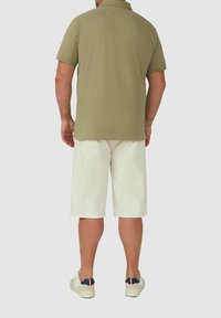 Mann trägt ein olivgrünes Poloshirt mit kurzen Ärmeln, weiße knielange Shorts und weiße Sneakers mit marineblauen und roten Akzenten, steht mit dem Rücken zur Kamera.