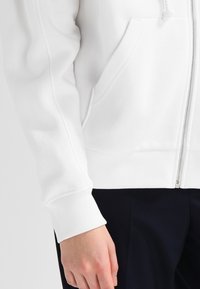 Hoodie zippé blanc avec une poche frontale, des poignets et un ourlet côtelés, une texture de tissu lisse et des détails de couture contrastés.