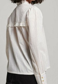 Mujer con una blusa blanca con sutiles rayas verticales, mangas largas abullonadas, detalle de volantes en el yugo trasero y botones dorados en los puños.