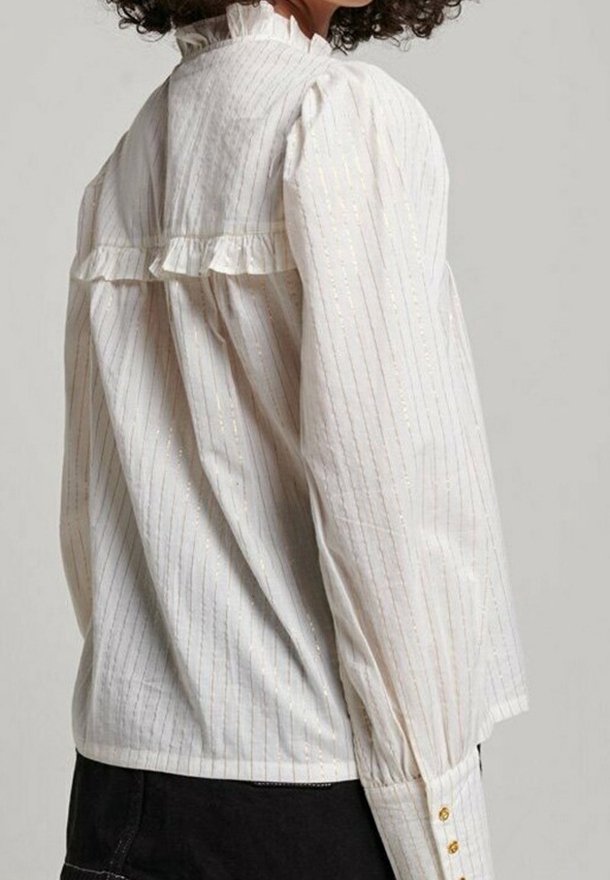 Mujer con una blusa blanca con sutiles rayas verticales, mangas largas abullonadas, detalle de volantes en el yugo trasero y botones dorados en los puños.