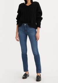 Suéter negro de punto trenzado, con cuello en V, de corte relajado. Jeans azules ajustados, de talle medio, con pierna cónica. Mocasines negros. Combinación de atuendo simple y casual.