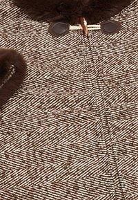 Brun och krämfärgad herringbone-mönstrad tyg med en texturerad finish, med en brun pälskrage och bruna läderknappar.