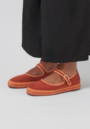 Ballerines Mary Jane en daim marron avec garniture beige et semelles en espadrille tissée, portées avec un pantalon noir à jambes larges.