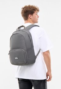 Gråtyg ryggsäck med ett huvudfack och en framficka, med vadderade remmar och ett handtag på toppen, bärs av en person i en vit skjorta.