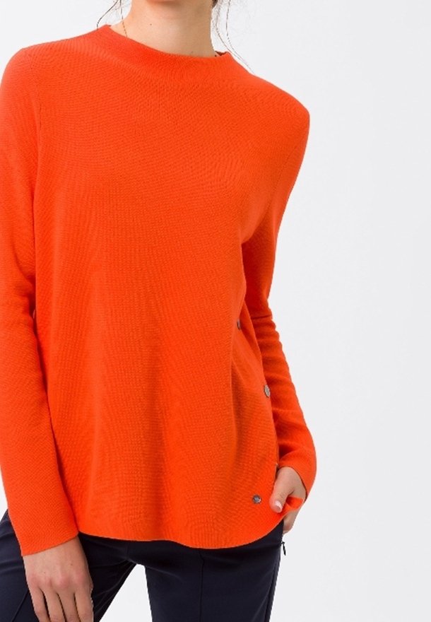 Femme portant un pull à manches longues orange vif avec des détails de boutons argentés sur le côté, associé à un pantalon foncé.