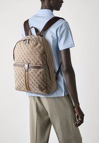 Sac à dos marron à motifs avec double fermeture éclair, porté sur le dos. Doté de bretelles marron foncé et d'accents beiges. Porté avec une chemise bleu clair.