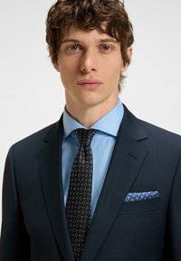 Marineblauer Anzugjacke mit strukturiertem Muster, hellblauem Hemd, schwarzer Krawatte mit geometrischem Muster und blauem Einstecktuch mit weißen Akzenten.