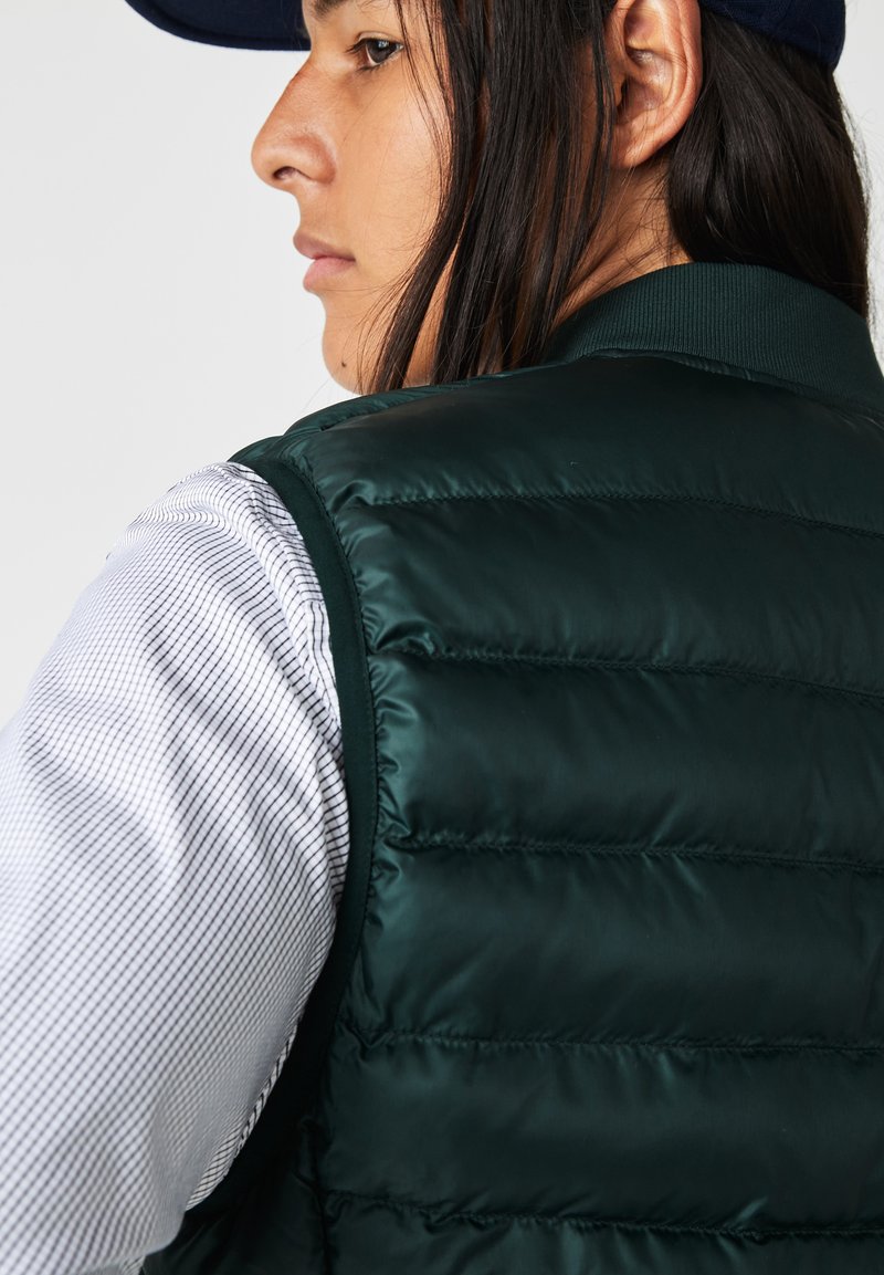 Chaleco de plumas verde con textura acolchada y cuello acanalado, llevado sobre una camisa de cuadros blanca y azul marino. El chaleco tiene un acabado brillante.