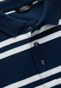 Chemise polo rayée bleu marine et blanche en tissu texturé avec un col, trois boutons noirs et une étiquette portant la mention "NEXT, FABRIQUÉ AU BANGLADESH."