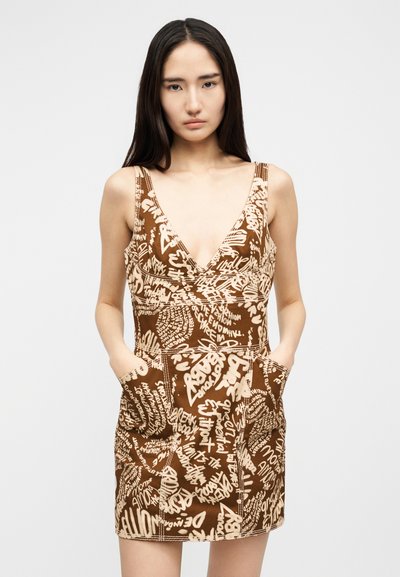 Νεαρή γυναίκα με μακριά σκούρα μαλλιά, που φοράει καφέ αμάνικο φόρεμα με μπεζ κείμενο σε στυλ γκράφιτι, χέρια στις μπροστινές τσέπες, ουδέτερο φόντο.