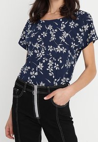 Námořnická blůza s bílým květinovým vzorem, krátké rukávy a kulatý výstřih. Kombinována s černými džíny s vysokým pasem a zipovým zapínáním.