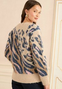 Gai bēšā džemperis ar pūkainu tekstūru, ar zilu, brūnu un rozā tīģera svītru rakstu, kā arī rievojumu aprocēm un apakšmalai.