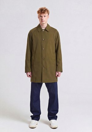 Junger Mann steht aufrecht, trägt einen olivgrünen Mantel mit Knöpfen, dunkelblaue Jeans und weiße Turnschuhe vor einem einfarbigen hellen Hintergrund.