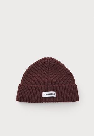 Un bonnet en tricot bourgogne avec une texture côtelée, présentant un revers et une étiquette rectangulaire blanche avec du texte noir.
