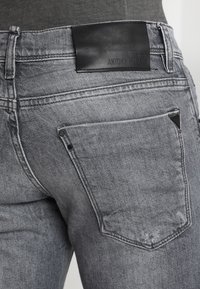 Jeans de mezclilla gris con un parche de logo de cuero negro, bolsillo trasero en ángulo con detalle de remache metálico y un acabado texturizado y desgastado.