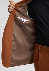 Brauner Außenstoffjacke mit einem glatten beigen Innenfutter, ausgestattet mit einer Tasche mit einem Knopfverschluss und einem Etikett, auf dem "feinster Stretchstoff" steht.