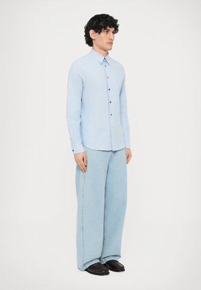 Cămașă albastru deschis cu nasturi multicolori, combinată cu blugi largi din denim deschis la culoare și pantofi negri, stând pe un fundal alb.