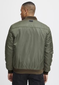 Olive green bomber jacket with a smooth, shiny texture, ribbed brown cuffs and hem, and a label on the back. Simple design, no additional details.