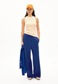 Gestreifter Tanktop in Weiß und Orange, blaue weite Hose, orange Kappe, hält einen blauen Cardigan, glattes Material mit lockerer Passform.