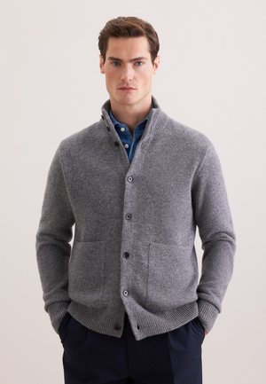 Mężczyzna ubrany w szary rozpinany sweter na guziki na niebieską koszulę, z rękami w ciemnych kieszeniach spodni, na jednolitym jasnym tle.