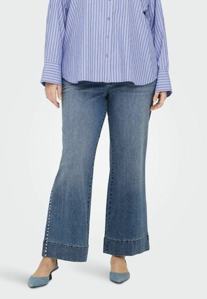 Jean large bleu avec un délavage moyen. Présente des rivets le long des coutures latérales et un ourlet retroussé. Porte avec une chemise à rayures à boutons.