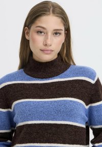 Maglione a righe con colletto alto, realizzato in morbido tessuto a maglia. I colori includono marrone, blu chiaro e bianco disposti in righe orizzontali.