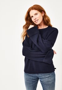 Dunkelblauer Strickpullover mit breiten Bündchen und strukturiertem Design. Kombiniert mit hellblauen Jeans, vor einem schlichten weißen Hintergrund präsentiert.