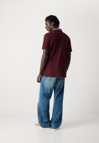 Man in een bordeauxrood polo t-shirt met korte mouwen, losse blauwe jeans met een scheur, en beige schoenen, staand met zijn gezicht naar rechts tegen een witte achtergrond.