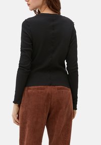 Musta pitkähihaine paita, jossa on aaltoileva reuna saumoissa, yhdistettynä ruskeisiin corduroy-housuihin, joissa on vaakasuoraa uritusta ja taskut.