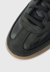 Melna ādas sporta apavu ar teksturētu virsmu, ar gludu priekšpusi, plakaniem auklām un gumijas zoli. Tīras šūšanas detaļas gar malām.