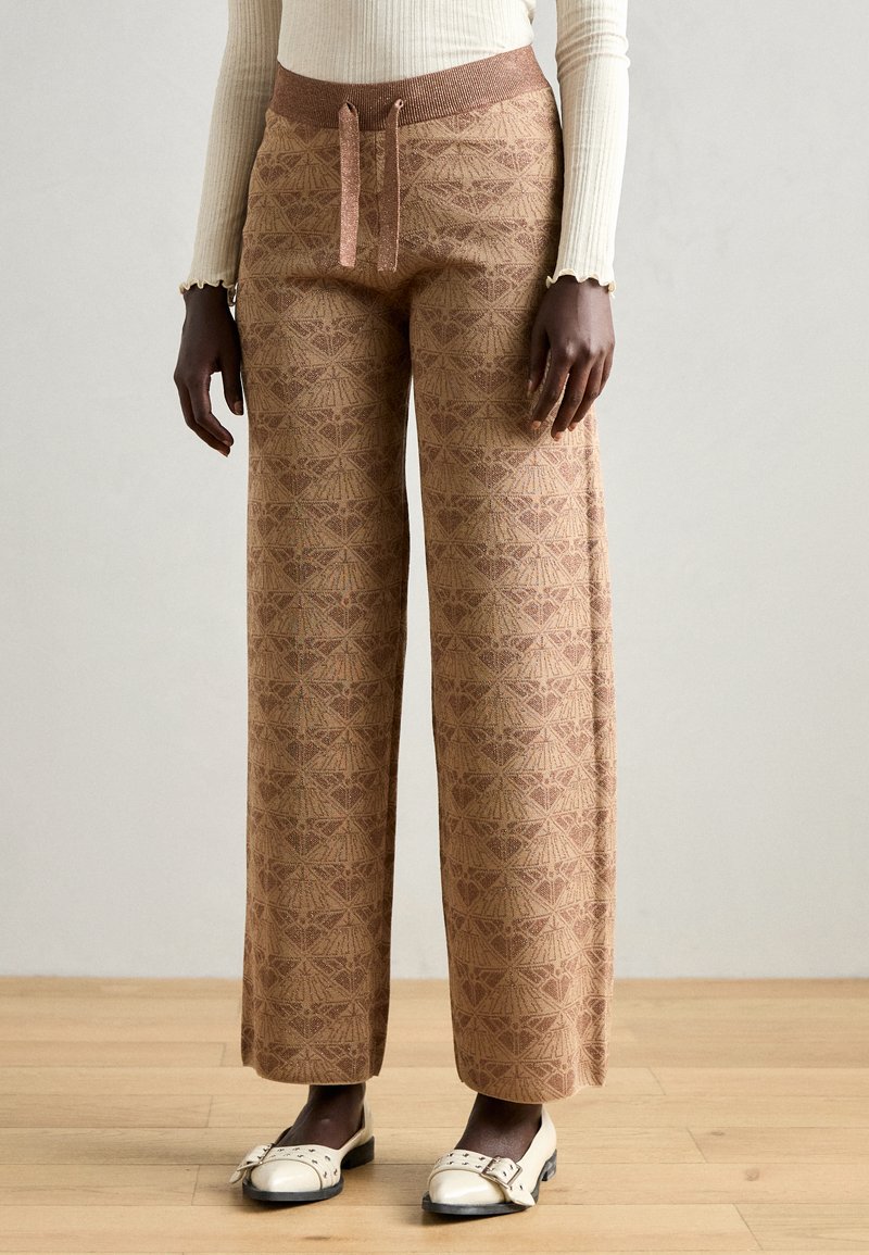 Pantalons larges à motif marron avec un design texturé. Ils présentent une taille élastique en tricot et un cordon de serrage. Associés à des chaussures blanches.