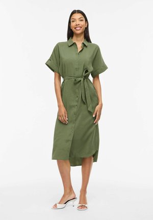 Lächelnde Frau in olivgrünem, knielangem Hemdkleid mit kurzen Ärmeln und gebundenem Taillengürtel, dazu weiße Mules mit Absatz, stehend vor weißem Hintergrund.