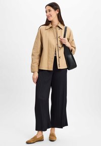 Beige leichte Jacke mit Knopfverschluss, kombiniert mit schwarzen plissierten weit geschnittenen Hosen und braunen Slippern. Schwarze Tasche mit Riemen.