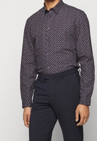 Langarm, donkerblauwe shirt met een bladenpatroon in wit en lichtblauw. Knopfafsluiting kraag, getailleerd ontwerp, katoenen stof, gecombineerd met donkere broek.