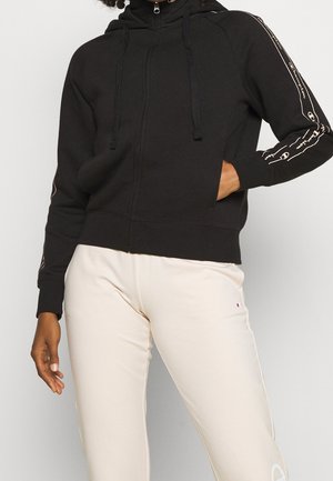 Sweat à capuche zippé noir avec un col haut et des cordons de serrage, présentant des détails de logo blanc le long des manches et un tissu doux et texturé.
