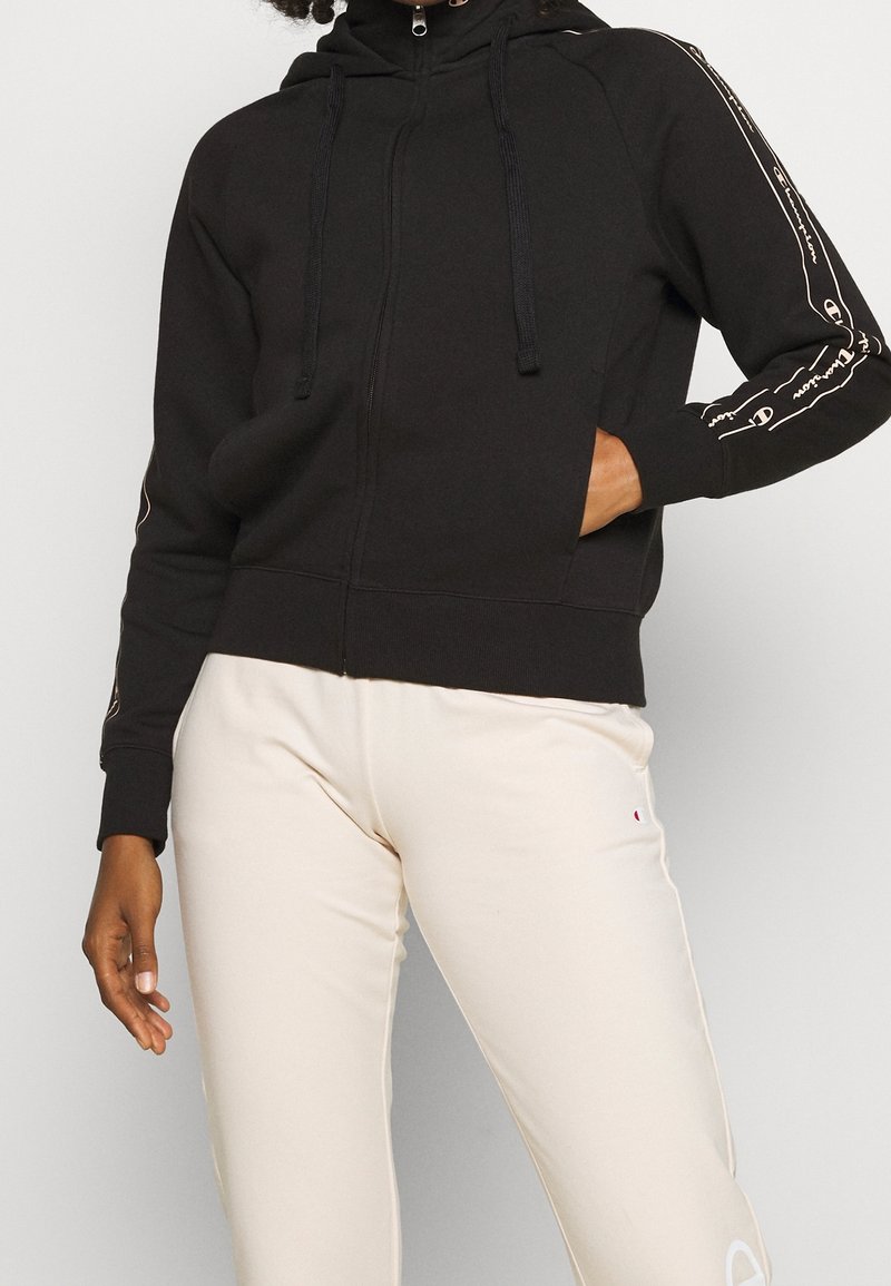 Felpa nera con zip e colletto alto, dotata di lacci, con dettagli del logo bianco lungo le maniche e un tessuto morbido e testurizzato.