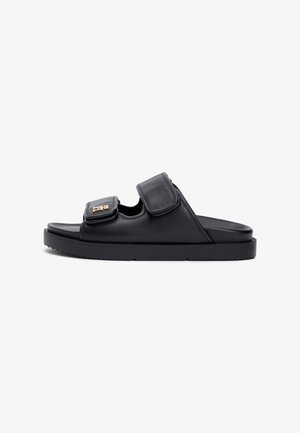 Sandales mules en cuir noir avec deux brides ajustables en velcro, un bout rond et une semelle épaisse en caoutchouc avec un motif de crampons texturé. Détails en métal doré.