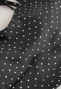 Tissu noir avec petits pois blancs, texture lisse et légères plis, suggérant un matériau pour vêtement ou accessoire.